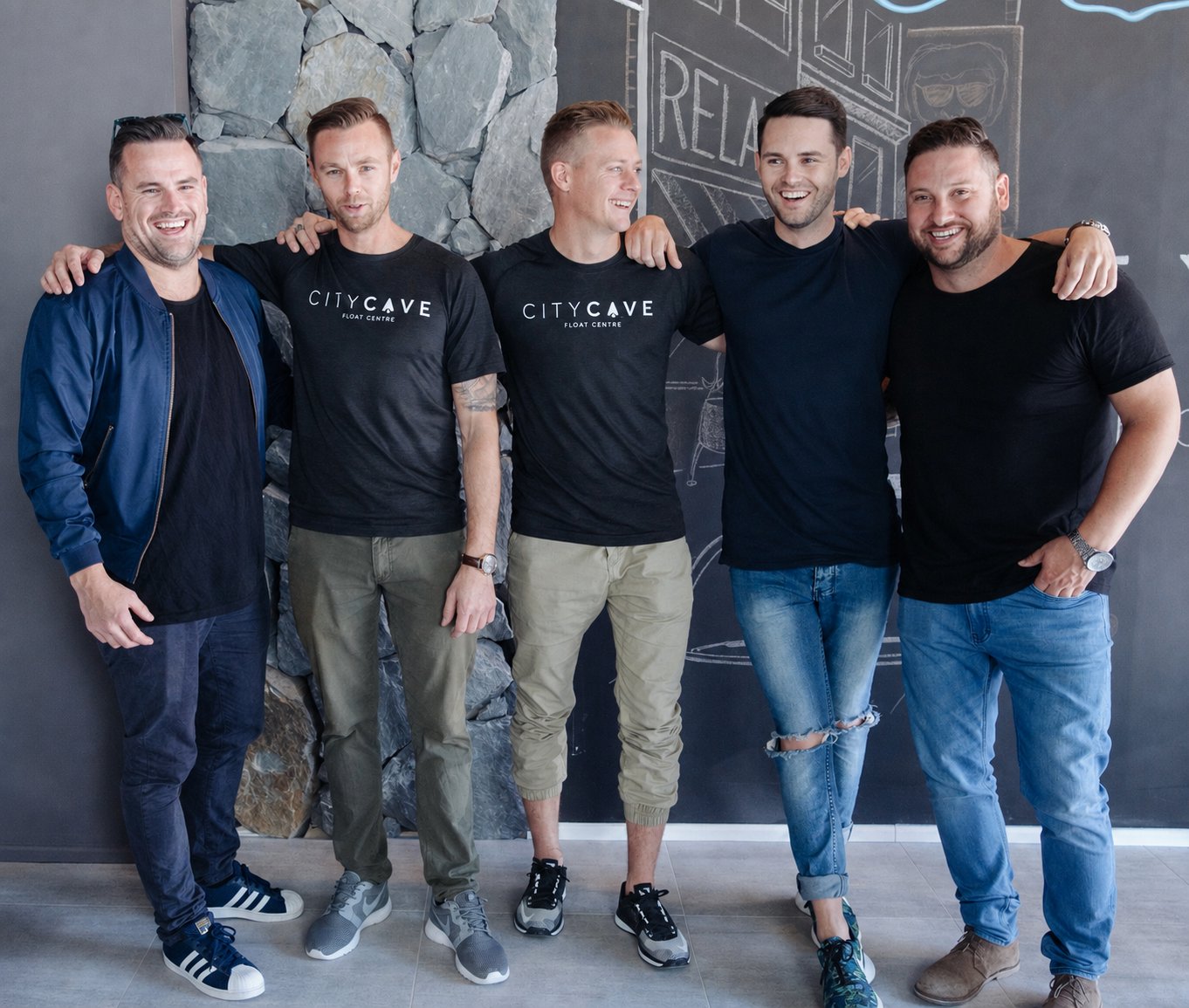 Five men side by side in front of the stone wall and chalk mural at the first City Cave centre, two wearing City Cave Float Centre t-shirts.