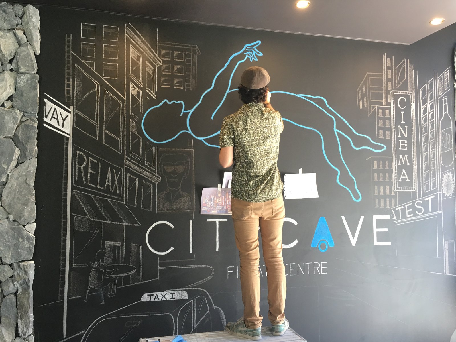 A front-on view of the chalk mural, the artist seen from behind, working on the blue floating figure above the hand-drawn City Cave wordmark.